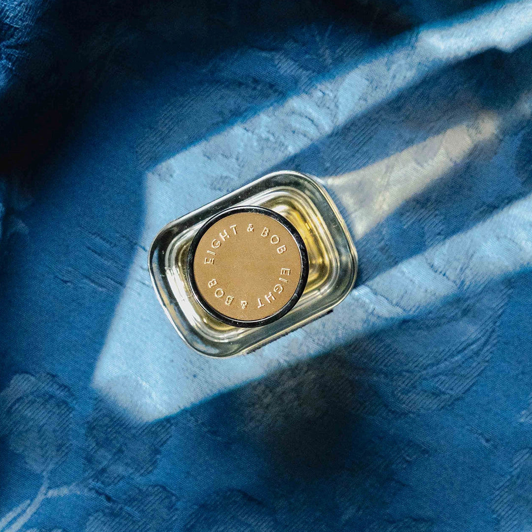 A small clear glass bottle with a gold-toned circular cap, placed on draped blue fabric with strong diagonal light and shadow.