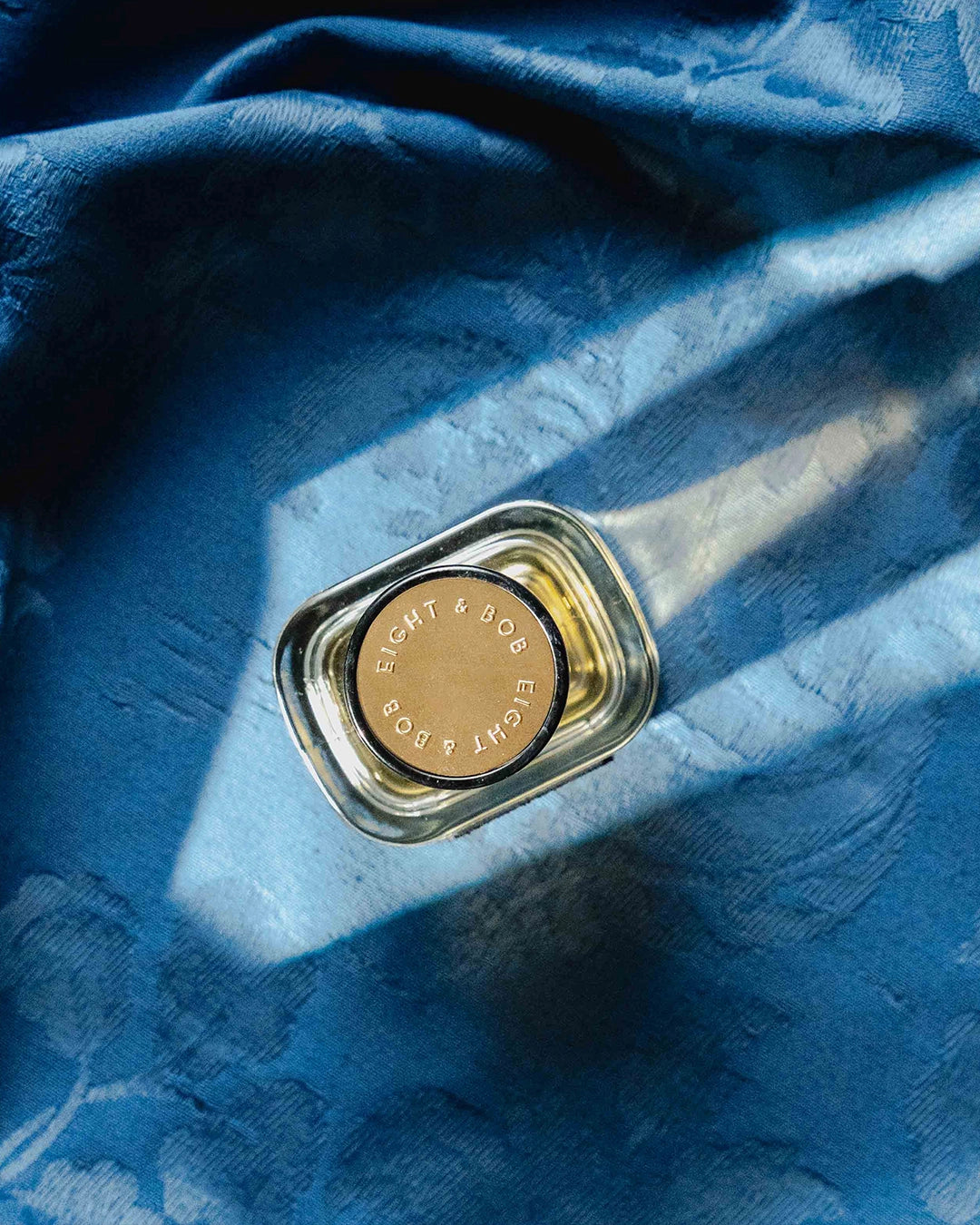 A small clear glass bottle with a gold-toned circular cap, placed on draped blue fabric with strong diagonal light and shadow.