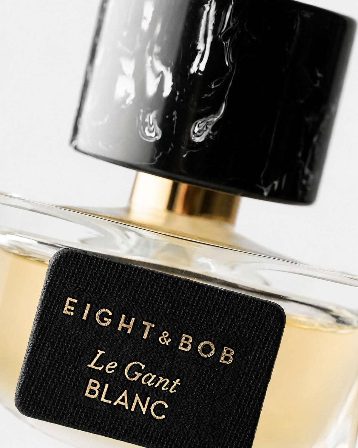 Close-up of an Eight & Bob Le Gant Blanc perfume bottle with a black textured cap, clear glass body, pale yellow liquid, and a black label with gold lettering.