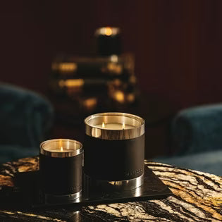 Two lit candles with black leather sleeves and silver rims sit on a tray on a marble table.