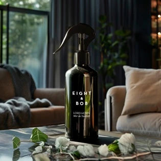 A black trigger-spray bottle labeled Eight & Bob sits on a dark reflective table in a living room with small white flowers.